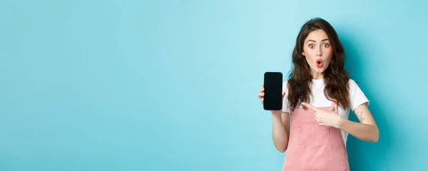 stock image Impressed young modern woman pointing at smartphone screen, say wow and stare amazed at camera, showing awesome application, new online store, standing over blue background.