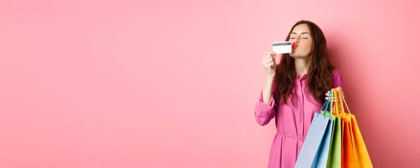 兴奋快乐的购物狂的画像 提着购物袋亲吻塑料信用卡的女人 惊讶的微笑 站在粉色的背景下 — 图库照片