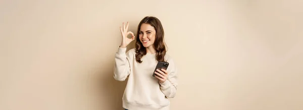 漂亮的女人手持手机 推荐应用程序 购物应用程序 站在米色背景之上 — 图库照片