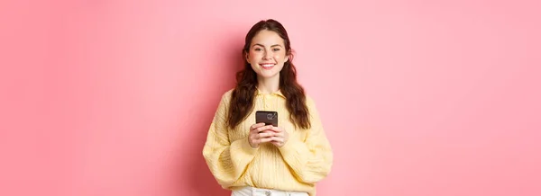 漂亮姑娘的画像 她手持手机 面带微笑 上网购物 用社交媒体应用程序 穿着休闲装站在粉色背景下 — 图库照片