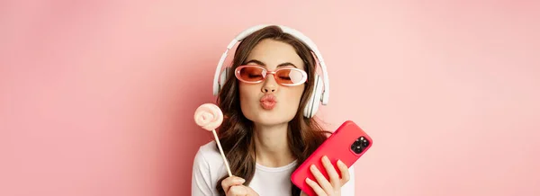漂亮的女模特戴着耳机听音乐 拿着棒棒糖和手机 戴着太阳镜 站在粉色的背景上 — 图库照片