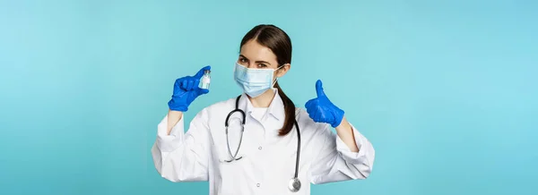 stock image Portrait of smiling doctor, medical personel in face medical mask and rubber gloves, showing thumbs up and coronavirus, omicron vaccine, standing over blue background.