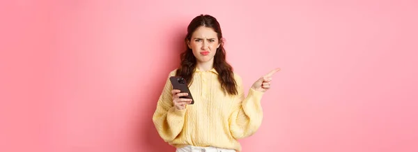 stock image Displeased girl grimacing, frowning upset and pointing finger aside at left copyspace while holding smartphone, standing over pink background.