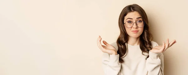 Stock image Young indecisive woman in glasses shrugging shoulders and looking confused, dont know, cant understand, standing over beige background.