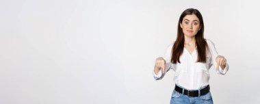 Doubtful cute girl shrugging unsure, smiling and pointing fingers down, standing over white background.