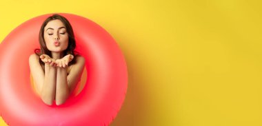 Portrait of beautiful female model posing with pink swimming beach ring, sending air kiss, mwah flirty, standing over yellow background.