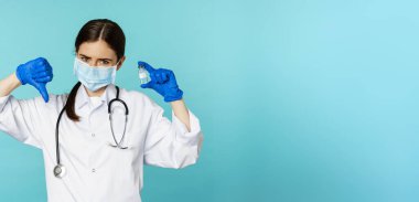 Üzgün ve hayal kırıklığına uğramış genç doktor, aşıyı gösteren ve sevmeyen kadın, jesti reddediyor, tıbbi ilacı onaylamıyor, mavi arka planda duruyor..