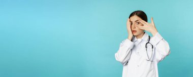 Shocked woman doctor, peeking through fingers, turn away from smth cringe ugly, standing in white coat over torquoise background.