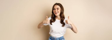 Excellent work. Happy young woman showing thumbs up in approval and smiling, standing in t-shrit over beige background.