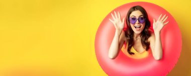 Close up portrait of enthusiastic young woman inside pink swimming ring, laughing and smiling, enjoying beach holiday, summer vacation, yellow background.