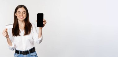 Portrait of female model showing credit card with smartphone screen, recommending application, standing over white background.