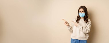 Health and covid-19 concept. Confused young woman in medical face mask, pointing left and staring with frustrated face, beige background.