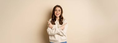 Happy smiling girl brunette pointing sideways, showing two promo offers, choice in store, standing over beige background.
