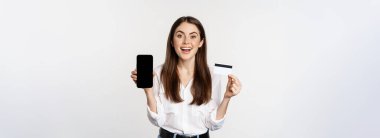 Happy woman showing credit card and smartphone screen, concept of online shopping, buying in app, standing over white background.