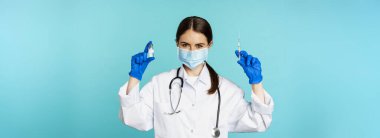 Gülümseyen sağlık personeli, maske takan doktor ve lastik eldiven, mavi arka planda duran covid-19 salgınından şırınga ve aşı gösteriyor..