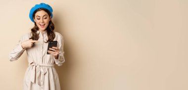 Portrait of stylish modern woman in outerwear, pointing at mobile phone screen and looking happy, smiling pleased, standing over beige background.