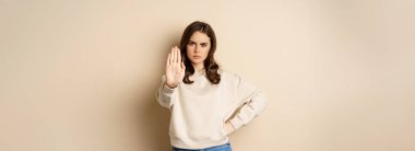 Stop. Serious and confident woman showing extended arm palm, prohibit, forbid smth, blocking something, standing over beige background.