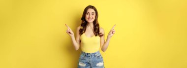 Portrait of stylish young female model, showing advertisement, pointing at logo or banner, demonstrating two ways, choices in store, standing over yellow background.