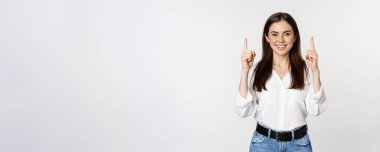 Smiling confident pretty woman, pointing fingers up, showing logo banner, announcement, standing in white blouse against studio background.