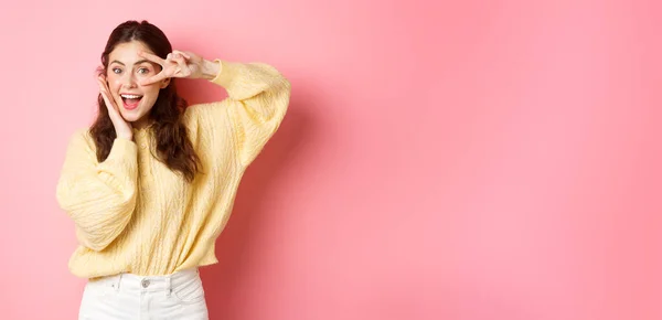 stock image Attractive female model, gasping amazed, showing v-sign over eye, smiling happy, standing against pink background.