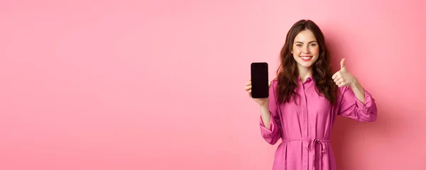 技术概念 穿着衣服的小女孩 展示着她的智能手机屏幕和大拇指 点头表示同意 称赞好的应用程序 站在粉色背景之上 — 图库照片