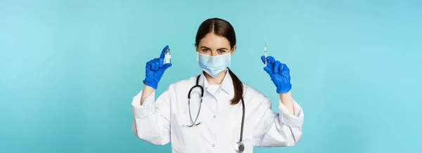 stock image Smiling medical staff, doctor in face mask and rubber gloves, showing syringe and vaccine from covid-19 pandemic, standing over blue background.