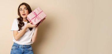 Celebration and holidays concept. Happy young girl looking intrigued, shaking box with gift, guessing whats inside wrapped present, standing over beige background.