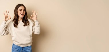 Very good, nice work. Cheerful young woman agrees, shows okay, ok praise, recommending smth, standing over beige background.
