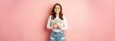 Loans and microcredit. Smiling beautiful girl showing money, cash in hands and looking enthusiastic, standing over pink background.