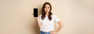 Online shopping and app concept. Young woman smiling and showing mobile phone screen, application interface, standing over beige background.