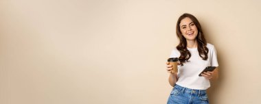 Smiling woman using mobile phone app and credit card, concept of online shopping, contactless payment and delivery, standing over beige background.