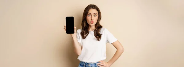 オンラインショッピングやアプリのコンセプト 若い女性は笑顔と携帯電話の画面を表示 アプリケーションインターフェイス ベージュの背景の上に立って — ストック写真