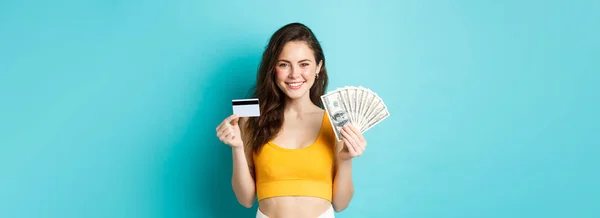 Stock image Portrait of young woman with smiling face, showing plastic credit card and money, standing against blue background.