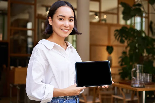 stock image Smiling asian woman showing digital tablet screen, cafe owner showing smth, standing in front of cafe entrance.