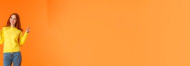 Dikey boyda, güzel, kızıl saçlı, kot pantolonlu, kışlık sarı süveterli, turuncu arka planda barış işaretleri olan, güler yüzlü, tasasız, neşeli, neşeli bir kız..