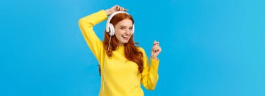 Yüzsüz, neşeli, mutlu, gülümseyen kızıl saçlı mutlu kadın eğleniyor, müzik dinliyor, ellerini kaldırıyor, dans ediyor, muhteşem ses kalitesinin tadını çıkarıyor, mavi arka planda dikilerek eğleniyor..