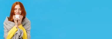 Bir kız sıcak kahvenin tadını çıkarıyor. Kapalı gözlerle aroma kokulu ve mutlu bir gülümseme. Sıcak gri eşarbı sarıp, kış sıcağında ısınıyor, mavi arka plan..