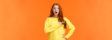 Dili tutulmuş ve etkilenmiş güzel, moda düşkünü, genç, modern, kızıl saçlı, düşük çeneli, nefes nefese kalmış, şaşırmış, kameraya bakan ve ilginç, harika bir ürünü işaret eden bir kız..