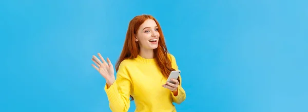 外向的女人 红头发 手持智能手机 给人发短信 挥手示意别人注意 打招呼 微笑打招呼 站在蓝色背景上 复制空间 — 图库照片