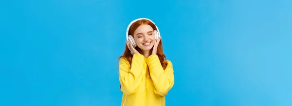 テクノロジー ガジェット ライフスタイルの概念 魅力的な屈託のない赤毛の女性は 大きな白いヘッドフォンで音楽を聞いて楽しむ 近い目とロマンチックな笑顔 立って青い背景 — ストック写真
