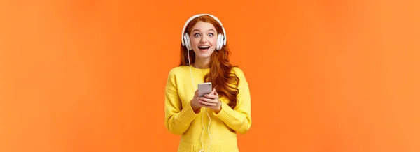 惊讶而又兴奋的红头发女孩发现她最喜欢的乐队发行了新歌 带着迷人而羡慕的眼神听着音乐 咧嘴笑着 拿着智能手机 戴着耳机 — 图库照片
