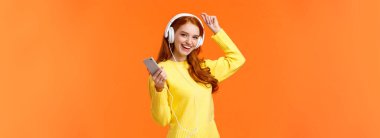 Hadi dans edelim. Neşeli, kaygısız, kıvırcık saçlı ve çilli modern hippi kız, dans eden, kaldıran, akıllı telefon tutan, yeni sipariş edilmiş online kulaklıklı, turuncu arka planda müzik dinleyen kız..