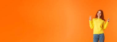 Dikey boyda, güzel, kızıl saçlı, kot pantolonlu, kışlık sarı süveterli, turuncu arka planda barış işaretleri olan, güler yüzlü, tasasız, neşeli, neşeli bir kız..