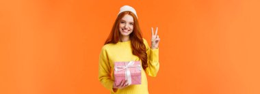 Kutlama, tatiller ve hediye konsepti. Kışlık şapkalı, kazaklı, elinde pembe bir hediye olarak barış işareti olan sevimli, neşeli, kızıl saçlı bir kadın, turuncu arka plan..