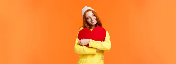 冬の帽子 セーター 愛の象徴として大きなかわいい赤いハートサインを受け入れ 楽しく笑顔 バレンタインデー オレンジの背景を祝う — ストック写真