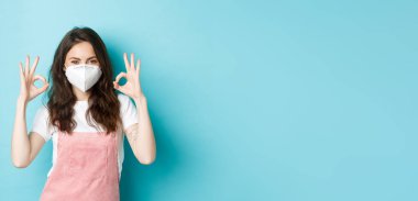 Covid, sağlık ve salgın hastalık konsepti. Solunum cihazında tatmin olmuş güzel bir kız, tıbbi maske onaylama işareti, koronavirüsten önleyici önlemler, mavi arka plan..