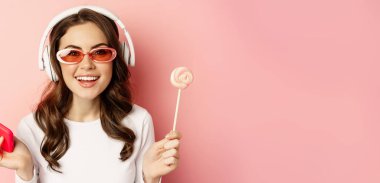 Close up portrait of stylish glamour girl, beautiful woman in headphones, listening music with smartphone, wearing sunglasses and licking lolipop.