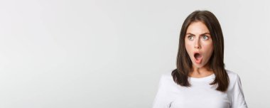 Etkilenmiş güzel kızın portresi sol tarafa bakıyor, çenesini indiriyor ve soluk soluğa kalıyor, şaşırıyor, beyaz arka plan.