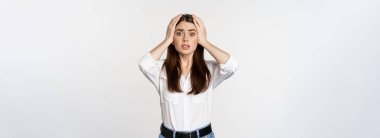 Worried and shocked young woman, looking stunned at disaster, holding hands on head, panicking, standing over white background.