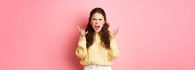 Angry and pissed-off girl screaming, shaking hands and grimacing furious, stare with outraged annoyed face at camera, standing against pink background.